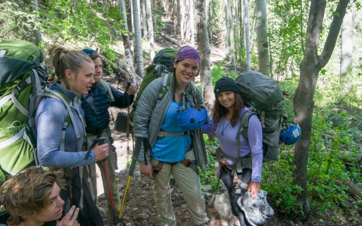 A group of students wearing backpacks and smile while standing in a shady area with green trees. 